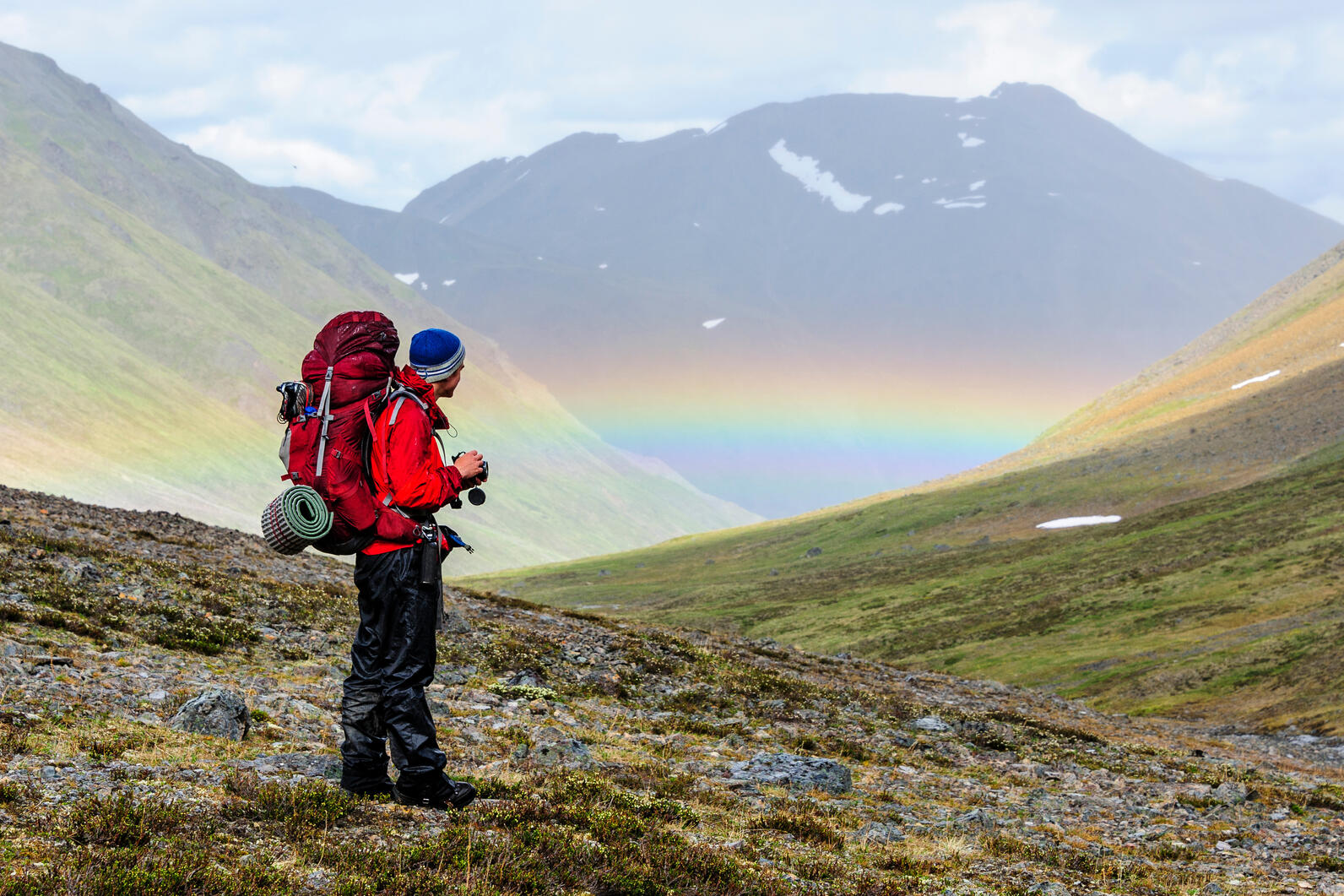 Man standing before green mountains and rainbow